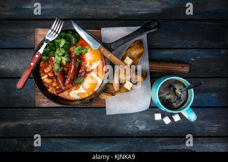 Frühstück in einer heißen Pfanne mit Spiegelei, Würstchen, Bohnen, Grün und Toasts. mit Kaffee rafinated Zucker und Besteck. top View Stockfoto