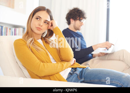 Schuss Riverside-flügel junge Frau in der Couch sitzen, während jungen Mann mit Laptop im Hintergrund. Stockfoto