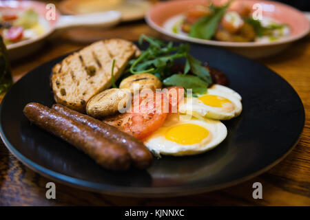 Enger Fokus auf Schüssel, amerikanisches Frühstück mit gegrillten Würstchen, Spiegeleier, Toast, Pilzen und Tomaten in Russischen Restaurant. Stockfoto