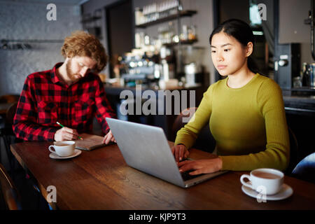 Asiatische Mädchen mit Laptop Vernetzung im Cafe mit ihrem Freund oder groupmate Notizen oder Skizzen in Notepad in der Nähe von Stockfoto