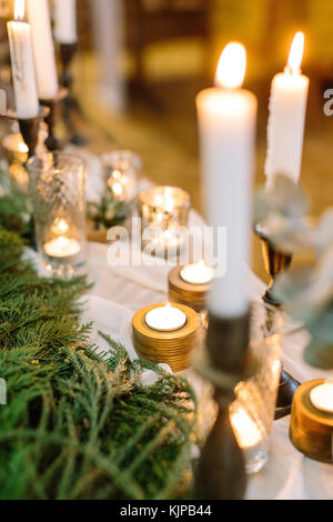 Beleuchtung, Komfort, Weihnachtsfest Konzept. Tabelle mit einer Vielzahl von Kerzen in verschiedenen Inhabern aus Glas, Gold, Holz, alle von ihnen auf der Kante, die durch frische fuzzy Koniferen Filialen platziert Stockfoto