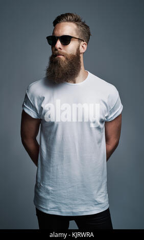 Stilvolle junger Mann mit langem Bart trug ein weißes T-Shirt und Sonnenbrille vor grauem Hintergrund stehend Stockfoto