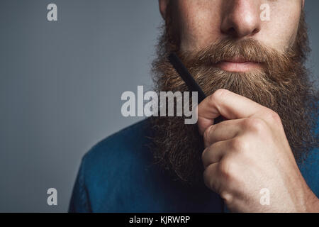 Nahaufnahme eines jungen Mannes styling seinem langen Bart mit einem Kamm, während allein in einem Studio auf grauem Hintergrund Stockfoto