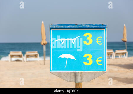 Blaue Schild für den Preis der Liegestühle und Sonnenschirme am Strand mit Strand und Geschirr in der Rückseite Stockfoto