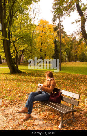 Frau sitzt auf einer Parkbank in der Stadt Parks und Wäldern im Herbst mit den Bäumen ihre Herbst Farben angezeigt. Stockfoto