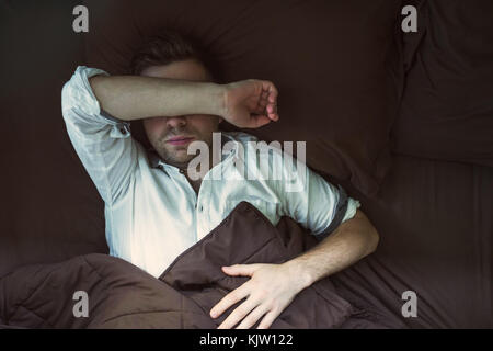 Portrait einer jungen kaukasischen Mann von oben schlafen in einem dunklen Bett. Er nach der Arbeit und Schlafen im weißen Hemd ist müde Stockfoto