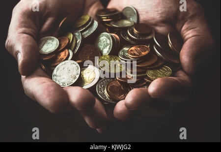 Nahaufnahme der hohlen Hände mit verschiedenen und bunten (Silber, Gold-, kupferfarben) Münzen auf einem dunklen Hintergrund. Stockfoto