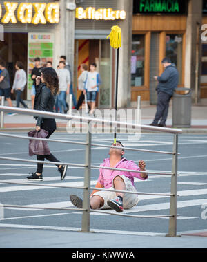 Eine Street Performer sitzen auf einem fußgängerüberweg in Bilbao Nordspanien Balancing eine Etage mop auf seiner Nase beim Rauchen einer Zigarette Stockfoto