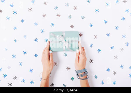 Weibliche Hände halten ein blaues Feld mit einem Geschenk unter dekorative Schneeflocken auf einem weißen Tisch Ansicht von oben Stockfoto