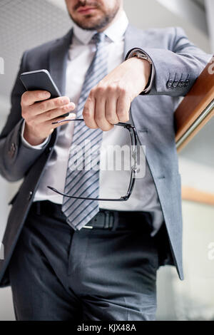 Nahaufnahme der unkenntlich Unternehmer mit Brille in der Hand stehen im geräumigen Büro Lobby und texting mit Kollegen auf dem Smartphone Stockfoto