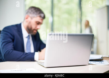 Besetzt Großraumbüro: Zuversichtlich in mittleren Jahren Manager im Sitzen arbeiten am Schreibtisch gewickelt, während seine Kollegin mit Panoramafenster Stockfoto