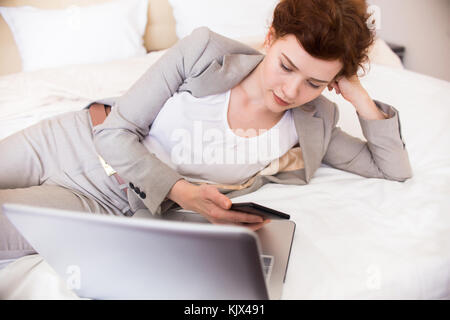 Portrait von hübschen jungen Geschäftsfrau Handauflegen hotel Bett ihr Telefon prüfen und mit Laptop und genießen Sie einen Aufenthalt in Business Travel Stockfoto