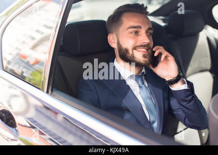 Portrait von gut aussehenden bärtigen Geschäftsmann sprechen am Telefon sitzen im Rücksitz des Autos und glücklich lächelnd Stockfoto