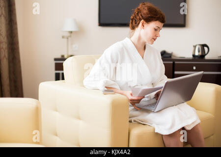 Seitenansicht Portrait von eleganten jungen Geschäftsfrau in Bademantel arbeiten mit Laptop und Dokumente im Hotel Zimmer während Geschäftsreisen gekleidet, kopieren Spa Stockfoto