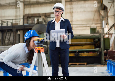 Nachdenklich Building Surveyor Notizen zu den Messungen, die während der Arbeit mit Ingenieurin durch optische Theodolit in der Factory Stockfoto
