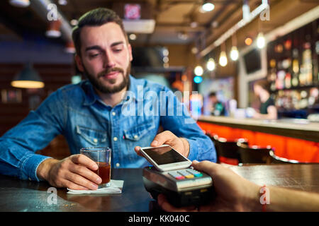 Portrait von betrunkenen jungen Mann bezahlen per Smartphone kaufen Getränke in der Bar, den Schwerpunkt auf die Payment Terminal im Vordergrund Stockfoto