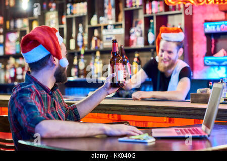 Portrait von bärtigen Barkeeper Tragen santa hut Jubel an Kunden mit Bierflasche in Pub, feiern Weihnachten, Fokus auf den Vordergrund Stockfoto