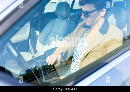 Portrait des erfolgreichen modernen Unternehmer tragen Sonnenbrillen, mit Laptop im Auto sitzen, Ansicht von hinter Glas Fenster Stockfoto