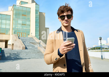 Portrait von hübscher junger Geschäftsmann Sonnenbrille tragen Sie ihr Smartphone stehen im Freien gegen moderne Bürogebäude, kopieren Raum Stockfoto