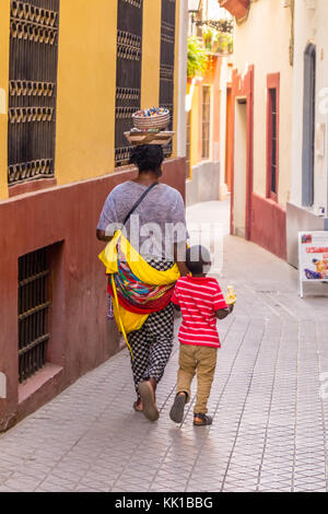 Afrikanischer Schmuckhändler, der ihre Waren in einem Korb auf dem Kopf trägt, und ihr Sohn in einer Seitenstraße, Sevilla, Andalusien, Spanien. Kind, das eine Banane isst Stockfoto