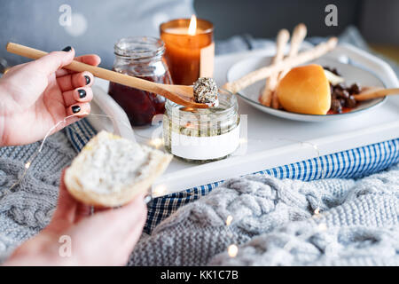 Weibliche Hände verschmiert Frischkäse mit frischem Brot. Frühstück im Bett, einem Tablett mit Käse, grissini, Marmelade und aromatische Kerze. Weihnachten Morgen. Honeymo Stockfoto