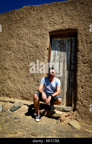 Kaukasische Mann mit T-Shirt und Jeans Shorts Hocken vor der alten Tür von Schlamm Haus Stockfoto