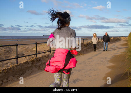 Frau, die an der Strandpromenade in Crosby, Merseyside, läuft. Wetter in Großbritannien. November 2017. Hell und sonnig an der Westküste der Mersey-Mündung. Dieses Küstengebiet um den Crosby Beach und den Crosby Marine Park erstreckt sich von Waterloo bis zur Mündung des Flusses Alt in Hightown. Der Strand und die Dünen sind derzeit die Heimat von 100 menschlichen Metallskulpturen, Antony Gormleys Another Place. Es gibt gefährliche Treibsand, so dass Besucher an der Küste empfohlen werden, sich innerhalb von 50 Metern von der Promenade zu halten. Kredit; MediaWorldImages/AlamyLiveNews Stockfoto