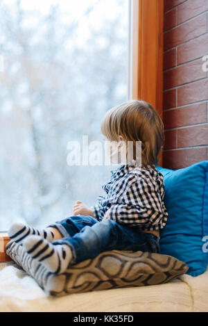 Little Boy In kariertes Hemd sitzt auf Kissen auf der Fensterbank Stockfoto