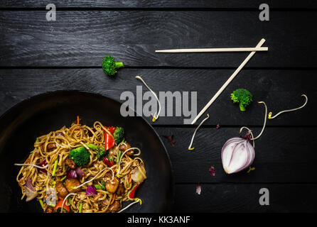 Udon fry Nudeln mit Huhn und Gemüse im Wok rühren Pan auf schwarzem Hintergrund mit Essstäbchen aus Holz. top View Stockfoto