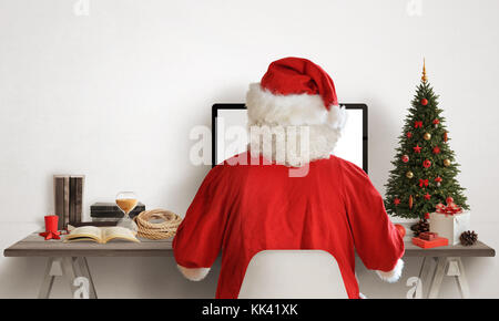 Santa claus Arbeit auf seinem Computer. Weihnachtsbaum und Geschenke neben. canta Schreibtisch mit Bücher, Karte, Seil. freier Platz für Text auf weißen Wand. Stockfoto