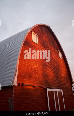 Schrägansicht der großen roten hölzernen Scheune mit gewölbten graues Metall Dach mit verstreuten Sonnenlicht und graue Wolken am Himmel überdachten Stockfoto