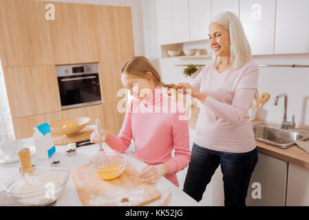 Schauen gut. angenehmen freudigen gealterte Frau steht hinter ihrer Enkelin und einen Zopf für ihr, während ihr zu helfen in der Küche Stockfoto