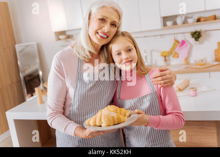 Home gemacht. Schön freut sich geschickte Großmutter und Enkelin stehen zusammen und lächelnd, während eine Platte mit Croissants Stockfoto