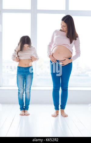 Adorable Ähnlichkeit. ansprechend süße Mädchen Prüfung ihren Bauch während der Schwangerschaft Mutter steht neben ihr und bewundern Sie, während Sie gegen Posing Stockfoto