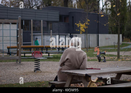 Großmutter auf einer Bank sitzen, spielen, beobachten die Kinder auf einem Spielplatz und einem gelben Fahrrad Stockfoto