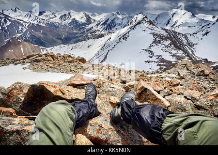 Die Beine des Menschen in Tracking Schuhe und Blick auf die verschneiten Berge bedeckt mit stürmischen Himmel Hintergrund Stockfoto