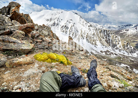 Die Beine des Menschen in Tracking Schuhe und Blick auf die verschneiten Berge mit bewölktem Himmel Hintergrund Stockfoto