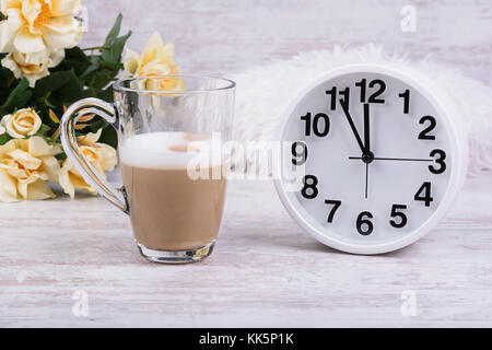 Hot latte Kaffee in klare Schale, weiß Wecker und schöne gelbe Rosen auf weiß Holz- Hintergrund. Stockfoto
