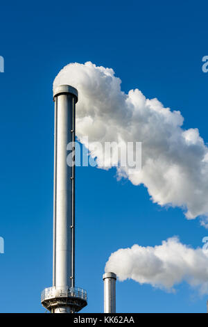 Zwei metallische Schornsteine spuckt eine dicke Wolke von weißem Rauch gegen den tiefblauen Himmel. Stockfoto