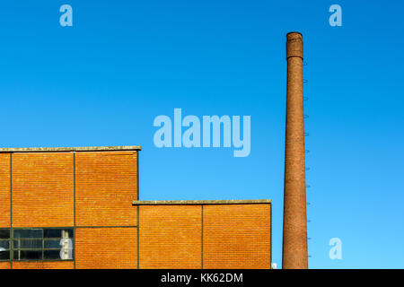 Der Backstein Fassade einer alten Fabrik mit einem hohen gemauerten Schornstein neben vor blauem Himmel. Stockfoto