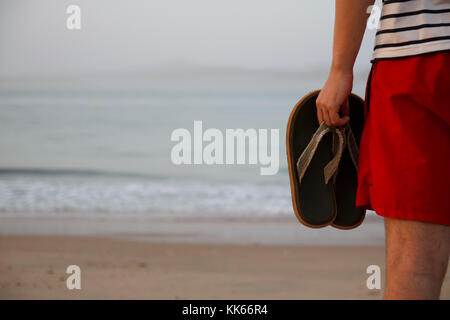 Mann am Strand bei Sonnenaufgang seine Sandalen Holding. Stockfoto