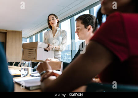 Frau mit einem Karton mit Kollegen am Tisch sitzen im Konferenzraum. Corporate Business Leute treffen im Board Room. Stockfoto