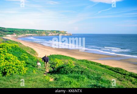 Mann zu Fuß Hund über Upgang Strand und Whitbys Dorf. Zwei Meilen westlich von Whitby an Nordseeküste in North Yorkshire, England Stockfoto