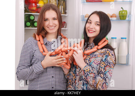 Zwei junge Mädchen hält Würstchen auf dem Kühlschrank Hintergrund. Zwei schöne junge Mädchen in der Nähe der Kühlschrank. Stockfoto