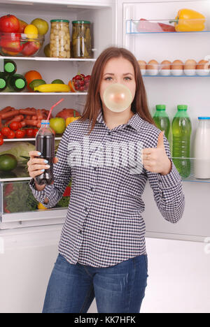 Junges Mädchen hält Cola und macht eine große Blase mit einem Kaugummi auf dem Kühlschrank Hintergrund Stockfoto