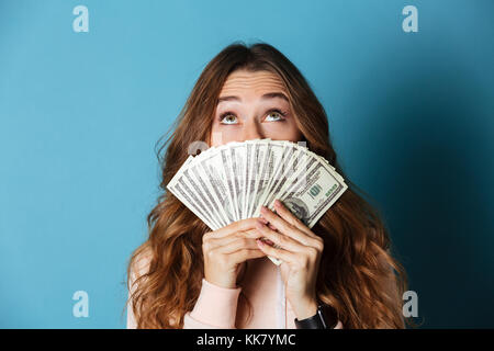 Portrait einer jungen hübschen Mädchen, die ihr Gesicht mit Bündel geld Banknoten und die Suche nach isolierten über blauer Hintergrund Stockfoto