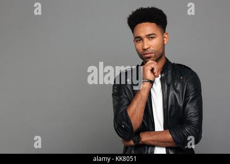Portrait einer jungen kontemplativen afro-amerikanische Mann in der Lederjacke stehend mit der Hand am Kinn und Kamera über grauer Hintergrund Stockfoto