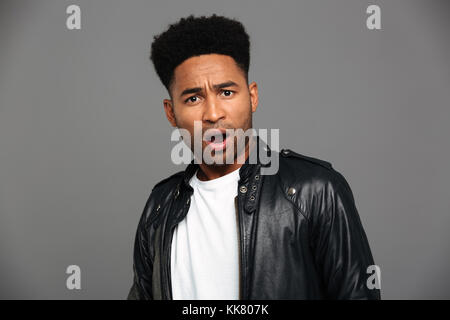 Porträt einer überrascht, schockiert afrikanischer Mann in Leder Jacke an Kamera über grauen Hintergrund suchen Stockfoto