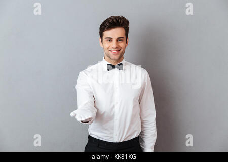 Porträt eines lächelnden freundliche Kellner in unifrom stehen mit ausgestreckten Hand über grauen Hintergrund gekleidet Stockfoto