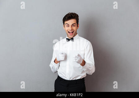 Porträt einer fröhlichen jungen männlichen Kellner in unifrom zeigen zwei Finger auf Kamera über grauen Hintergrund gekleidet Stockfoto